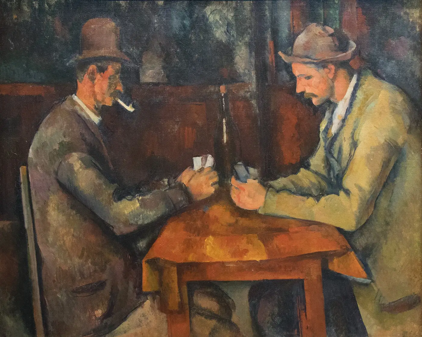 Two men in hats play cards across a small wooden table, one with a pipe.